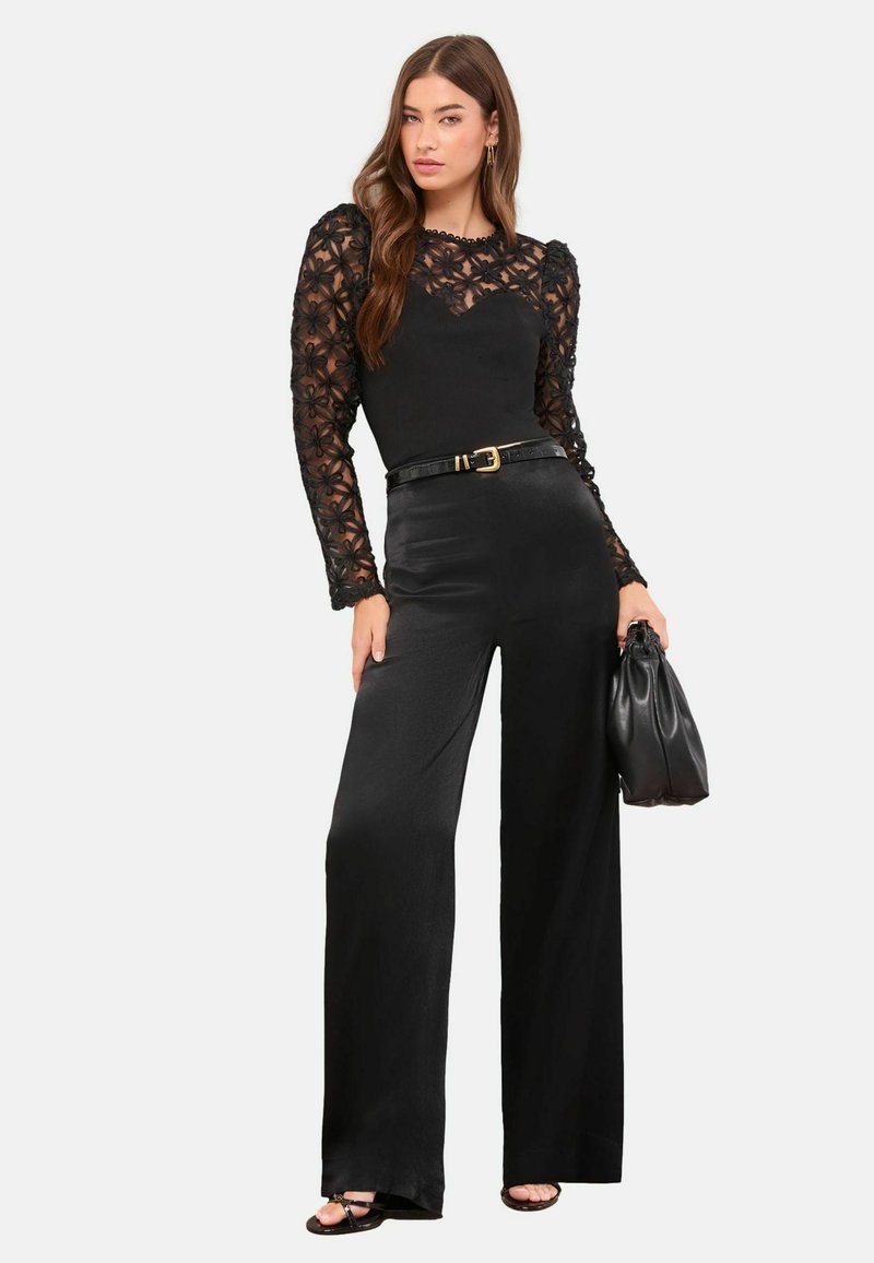 Juodas kombinezonas su ilgomis permatomomis nėrinių rankovėmis, puoštomis gėlių raštais. Šilkinio pojūčio audinys, plačios kelnės, liemuo pabrėžtas juodu diržu.