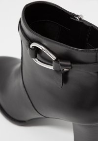Botín negro de cuero hasta el tobillo con una forma estrecha, que presenta una hebilla de metal plateado y un acabado texturizado en el borde superior.