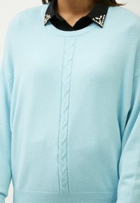 Hellblauer Pullover mit einem Zopfmuster in der Mitte, runder Ausschnitt und schwarzer Hemdkragen, verziert mit Strasssteinen.