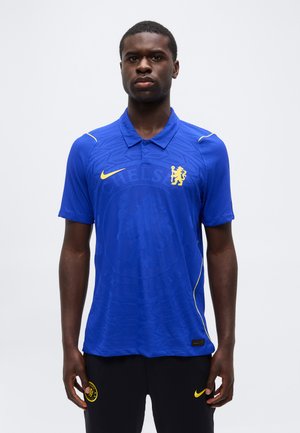 Mężczyzna ubrany w niebieską koszulkę piłkarską Chelsea z żółtym logo i czarne spodnie z żółwym znakiem Nike i emblematem Chelsea, stojący na jednolitym tle.