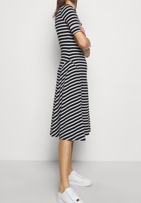 Woman standing with arms crossed wearing a black and white striped midi dress and white sneakers against a plain background.