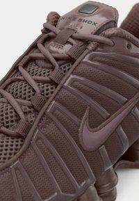 Nike Shox sneakers featuring a dark purple mesh upper, prominent rubber spikes, and a smooth plastic accent with laces woven through loops.