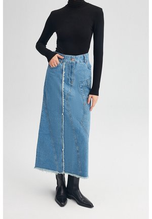 Femme portant une jupe midi en denim bleu bicolore à ourlet effiloché, un col roulé noir et des bottes noires à bout pointu, debout avec une main dans la poche.