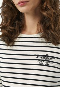 Gestreiftes Langarmhemd in Weiß und Schwarz mit einem gestickten Haifischdesign und dem Text "COMMA LOVES PORTUGAL" darunter. Weicher Stoff.