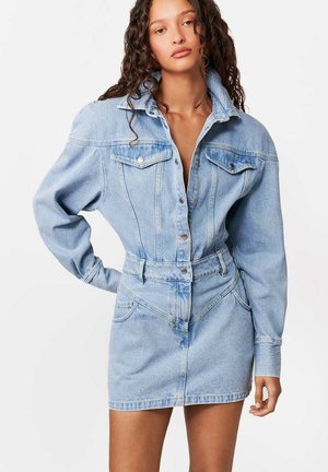 Kobieta o długich, kręconych włosach, ubrana w jasnoniebieską sukienkę dżinsową zapinaną na guziki, z bufkami na długich rękawach i przednimi kieszeniami, stojąca z jedną ręką na biodrze.