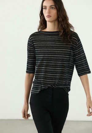 Femme aux longs cheveux bruns portant un haut à manches courtes rayé noir et blanc et un pantalon noir, debout devant un fond clair uni.