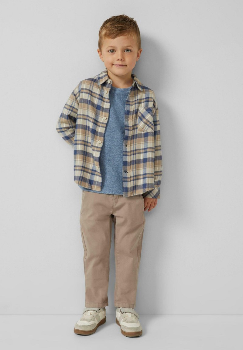 Jonge jongen die tegen een grijze muur staat, gekleed in een beige en blauw geruit overhemd, een blauwe T-shirt, beige broek en witte sneakers.