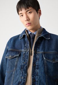 Jeansjacka i mörkblått med påtagliga sömmar, två bröstfickor, bärs över en beige zip-up-top och marinblå slips, mot en vit bakgrund.