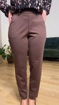 Donna che indossa pantaloni marroni sartoriali a vita alta e una camicetta nera a fantasia, in piedi al chiuso con le mani nelle tasche.