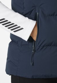Giacca puffer blu navy con tasca con zip, abbinata a una maglietta a maniche lunghe bianca con strisce diagonali nere sulle maniche.