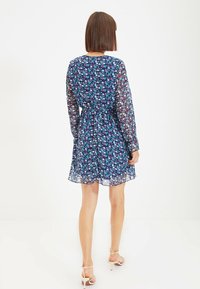 Kukallinen mekko tummansinisellä taustalla, jossa on pitkät läpikuultavat hihat, vyötäröltä kootut ja virtaava helma, jossa on pieniä, värikkäitä kukkakuvioita.