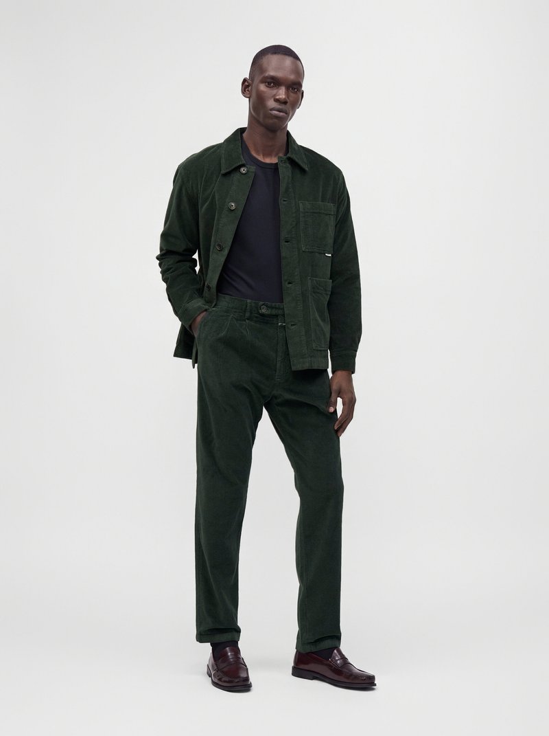 Homme debout portant une veste et un pantalon en velours côtelé vert foncé, une chemise noire et des chaussures en cuir marron foncé sur un fond clair uni.