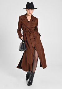 Brauner Wildleder-Trenchcoat mit einem Gürtel zum Binden und einem tiefen Schlitz. Die Accessoires umfassen eine schwarze Handtasche und spitze schwarze Stiefel.