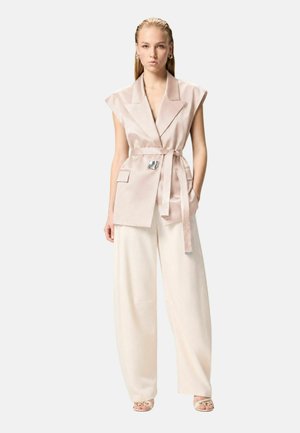 Donna che indossa un blazer beige senza maniche con cintura e grandi revers, pantaloni crema a gamba larga e sandali beige con dettaglio a catena.
