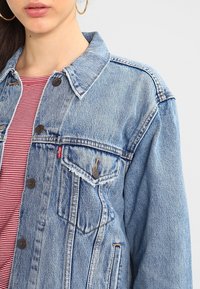 Blå jeansjacka med klassisk skärning, med krage, framfickor och silverknappar, kombinerad med en röd- och vitrandig skjorta.