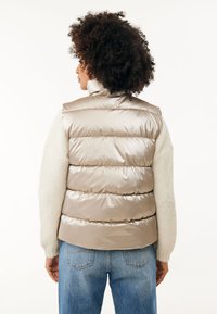 Beige satin väst med hög krage och horisontell quiltning, kombinerad med en ljus, texturerad tröja och blå jeans.