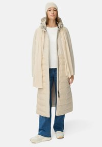 Kvinde iført en beige lang polstret frakke, matchende tørklæde og hue, hvid skjorte, blå jeans og hvide sneakers, der står foran en hvid baggrund.