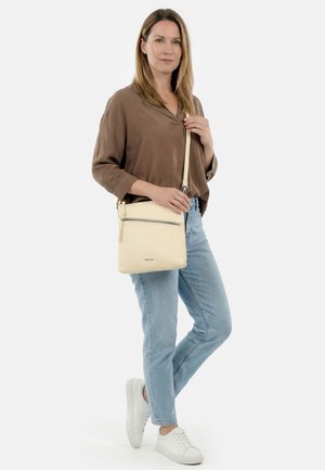 Femme debout avec un sac bandoulière beige clair, portant un chemisier marron, un jean bleu clair et des baskets blanches sur fond blanc.