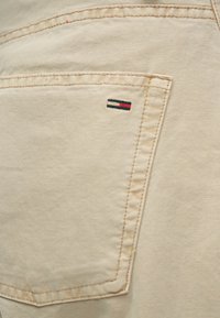 Smėlio spalvos audinys su tekstūruotu paviršiumi, turintis užpakalinę kišenę ir mažą išsiuvinėtą logotipą tamsiai mėlynos, baltos ir raudonos juostelėmis.