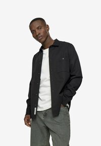 Fekete gombos ing gallérral, elülső zsebbel és hosszú ujjakkal, fehér pólóval és világosszürke nadrággal párosítva.