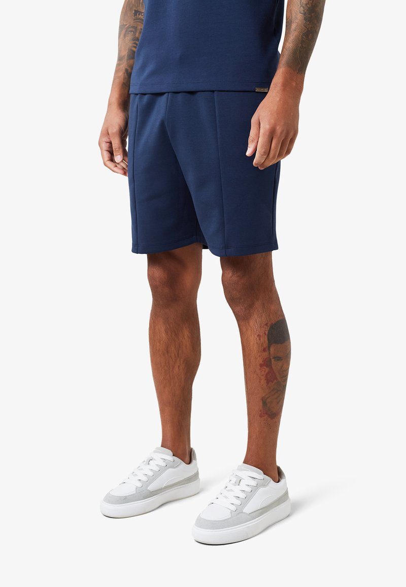 Homme portant un short bleu marine et une chemise assortie, des baskets blanches, avec des tatouages visibles sur les bras et le mollet droit.