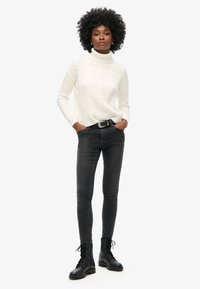 Witte kabelgebreide coltrui, slim-fit donkere jeans en zwarte veterschoenen. Gelaagd stof met verhoogde patronen en een getailleerde silhouette.