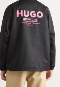 Czarna kurtka z różowym logo "HUGO" na plecach, z tekstem zapisanym pogrubioną czcionką, gładką strukturą i luźnym krojem.