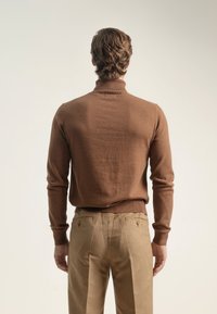 Hombre de pie de espaldas, vistiendo un suéter marrón de cuello alto y pantalones beige contra un fondo claro y liso.