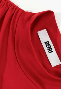 Rode stof met een gladde textuur, met verwerking rond de halslijn. Een label met "BENU" is zichtbaar, genaaid in het kledingstuk.