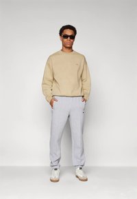 Bēša sporta džemperis, pelēkas sporta bikses, baltas sporta kurpes un melnas saulesbrilles. Brīvs piegriezums ar ribām aprocēs un smalku logotipu uz džempera.