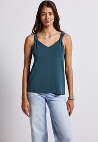 Teal V-halsad ärmless topp med plisserade axelband. Mjuk tyg, avslappnad passform och rak nederkant. I kombination med ljusblå vida jeans.