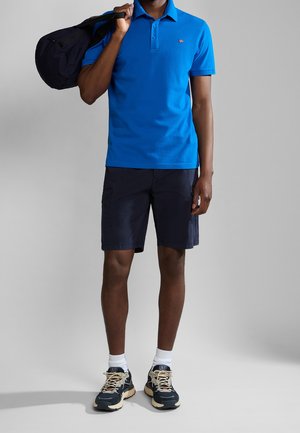 Homme portant un polo bleu vif, un short cargo foncé, des chaussettes blanches et des baskets noires, tenant un sac de sport noir sur l'épaule.