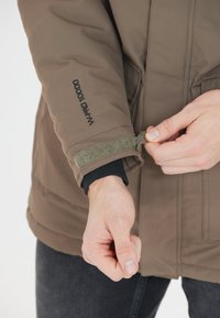 Brun isolerad jacka med en strukturerad yta, justerbart ärmslut och synlig logotyp "W-PRO 10000" på ärmen. Svart innerfoder synligt vid ärmslutet.