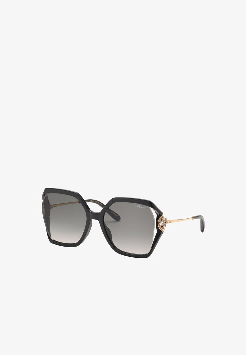 Lunettes de soleil hexagonales noires oversize avec verres dégradés gris et branches décoratives dorées ornées d'un petit emblème près des charnières.