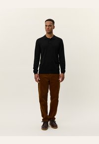 Zwarte langemouwen polo met kraag, gecombineerd met bruine corduroy broek en bruine schoenen, tegen een zachte achtergrond.