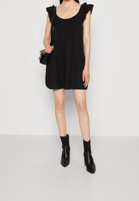 Vestido negro sin mangas con acentos de hombros con volantes, detalle de cintura fruncida y un corte holgado. Lleva botas de tobillo negras con tacón.