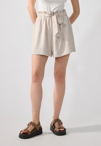 Shorts beige clair avec une taille élastique et un détail de lien, présentant un tissu texturé et une coupe ample, associés à des sandales ouvertes marron.