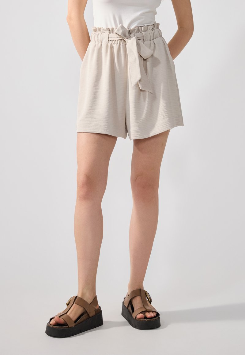 Shorts beige clair avec une taille élastique et un détail de lien, présentant un tissu texturé et une coupe ample, associés à des sandales ouvertes marron.