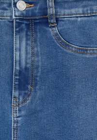 Blå denimtyg med en slät textur, med kontrasterande orange sömmar, en framficka och en metallknapp vid midjan.