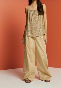 Femme portant un haut sans manches à motifs et un pantalon beige à jambes larges, pieds nus, se tenant contre un fond orange.