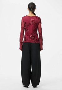 Burgunderfarget langermet topp med blomsterbroderi på gjennomsiktig stoff, kombinert med vide svarte bukser, som skaper en kontrasterende silhuett.