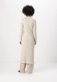 Robe tricotée en crème avec un tissu texturé, manches longues, longueur mi-mollet, se distinguant par un ourlet à franges et un détail de boutons dans le dos.