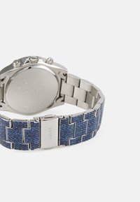 Reloj de acero inoxidable con una correa de patrón de mezclilla azul. Presenta un acabado pulido, detalles en plata y una caja redonda con diales de cronógrafo.