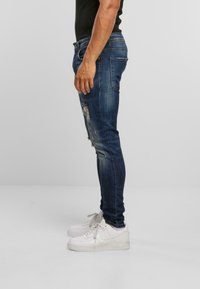 Muž v tmavě modrých roztrhaných skinny džínách a bílých teniskách stojící bokem na jednobarevném pozadí.