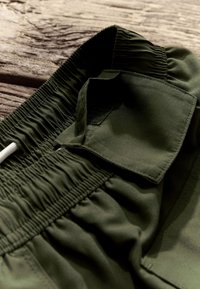 Tumedrohelised lühikesed püksid elastse vöökohaga, millel on takjakinnitusega taskukaas ja sile, kerge kangas. Äärte ümber tekstuuriga õmblused.