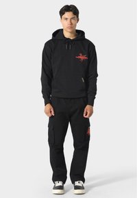 Sudadera negra con bolsillo tipo canguro y logo gráfico rojo. Combinada con pantalones cargo negros con bolsillos y un acabado texturizado.