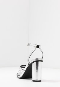 Srebrny sandał na wysokim obcasie z cienkimi paskami, metalicznym słupkiem i gładką czarną podeszwą. Posiada pasek na kostkę z klamerką.