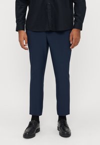 Marineblaue Hose mit maßgeschneiderter Passform, gefertigt aus glattem Stoff. Kombiniert mit einem schwarzen Hemd und schwarzen Schuhen, mit einem einfachen, eleganten Design.