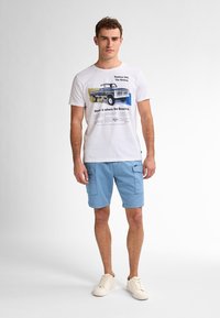 Biała koszulka bawełniana z grafiką samochodu i napisem, w połączeniu z jasnoniebieskimi szortami z kieszeniami i białymi sneakersami. Prosty design.