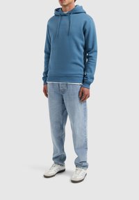 Blauwe hoodie gemaakt van een zachte katoenmix met een voorzak in kangoeroestijl. Gecombineerd met lichtblauwe jeans en witte sneakers met grijze accenten.